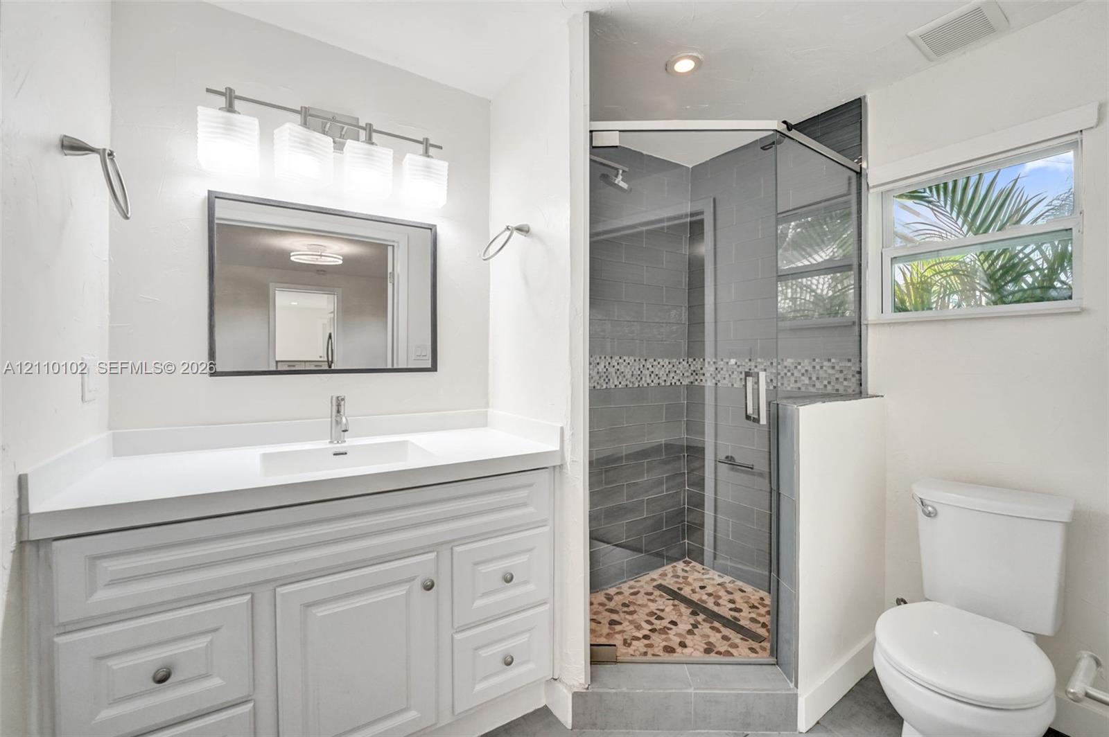 White cabinetry vanity with a clean-lined countertop, rectangular undermount sink, four-light vanity fixture, corner glass-enclosed shower featuring decorative tile accents and pebble-style flooring, and a single-hung window
