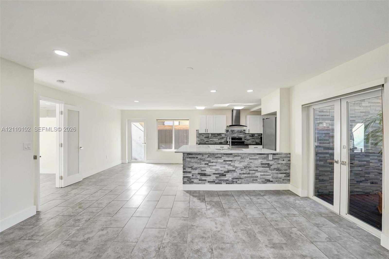 Open-concept interior featuring large-format ceramic tile flooring, kitchen island with stone-finish cladding, quartz countertops, stainless steel appliances, and French doors
