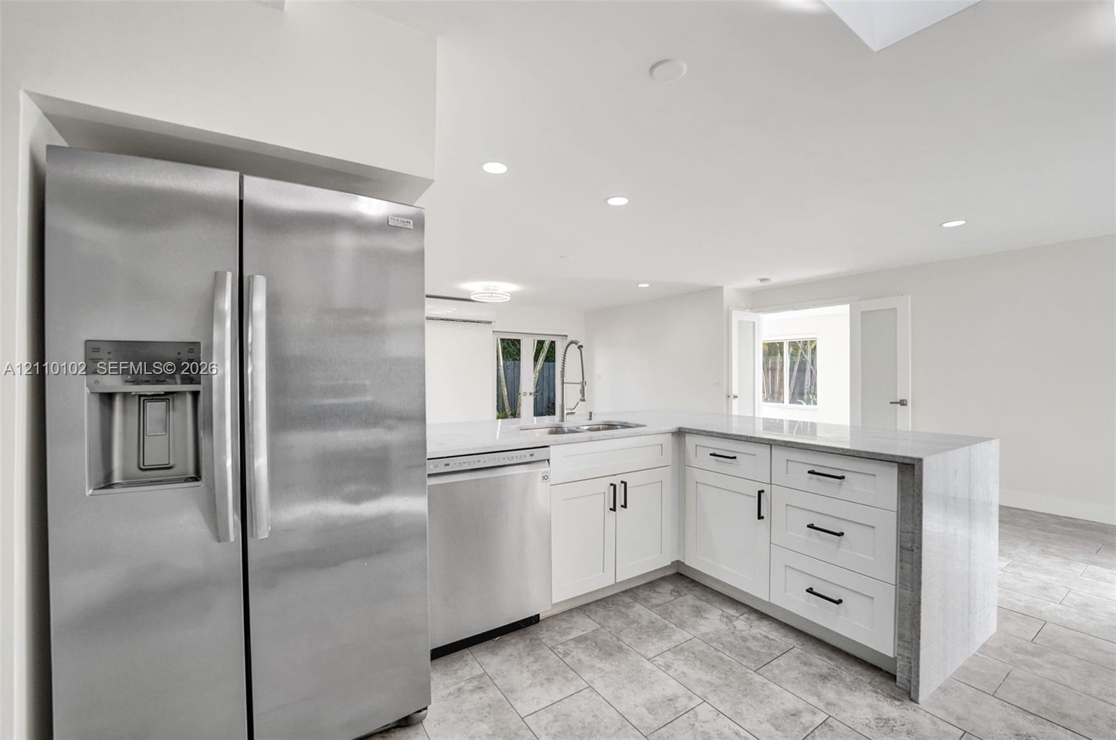 Kitchen featuring white shaker-style cabinetry, stainless steel appliances, quartz countertops, a waterfall-edge peninsula, and stone-tile flooring