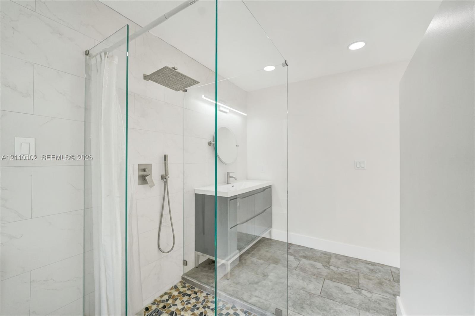Walk-in shower featuring a glass enclosure, rainfall showerhead, and wall-mounted hand sprayer, complemented by a dual-drawer vanity with a white countertop and recessed lighting