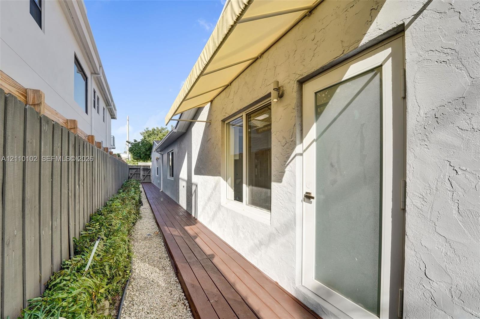Stucco exterior featuring a wood-finish deck, retractable fabric awning, frosted-glass entry door, and perimeter privacy fencing