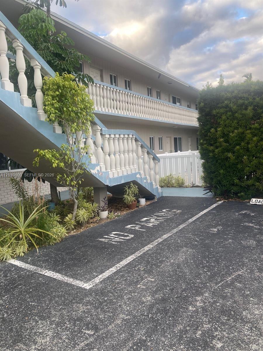 Exterior of multi-story building featuring painted concrete balustrade railings, exterior staircase, asphalt paved ground surface, and perimeter white vinyl fencing