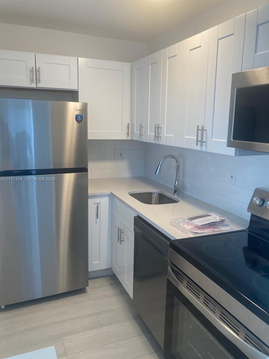 Kitchen featuring white shaker-style cabinetry, light-toned stone countertops, stainless steel appliances, a white tile backsplash, and wood-finish flooring