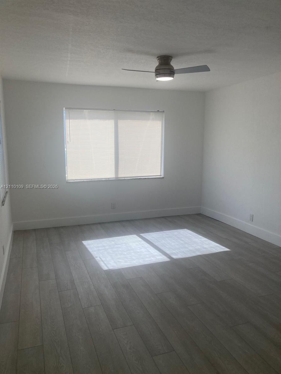 Neutral-toned interior room featuring wood-finish flooring, a ceiling-mounted light fixture with a fan, and a single window with fitted horizontal blinds