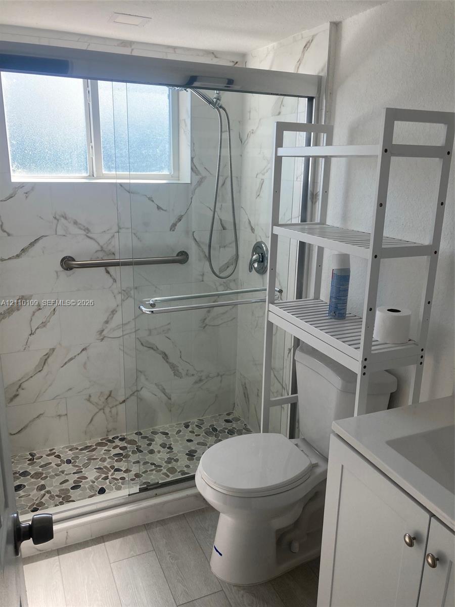 Walk-in shower featuring large-format tile surround with stone-pattern detail, pebble-texture shower floor, sliding glass door, and metal grab bar, complemented by wood-finish flooring