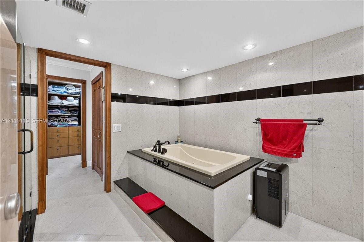 Soaking tub featuring a black stone surround and deck, recessed lighting, floor-to-ceiling wall tile with black accent inlay, and wood-finish built-in closet cabinetry