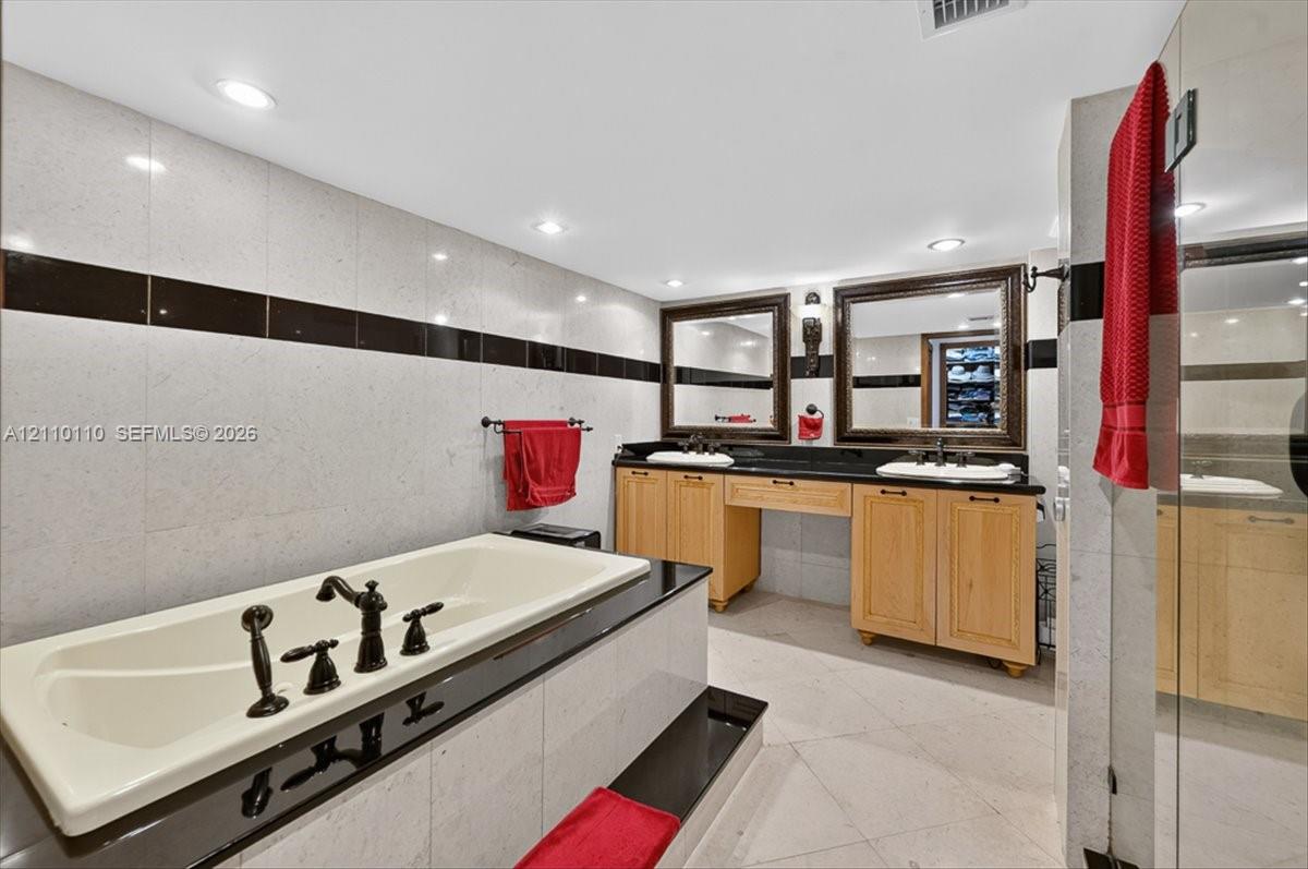 Dual-vanity cabinetry with light wood finish, integrated stone-surface countertop, recessed lighting, drop-in soaking tub with dark-finish hardware, and tile wall accents