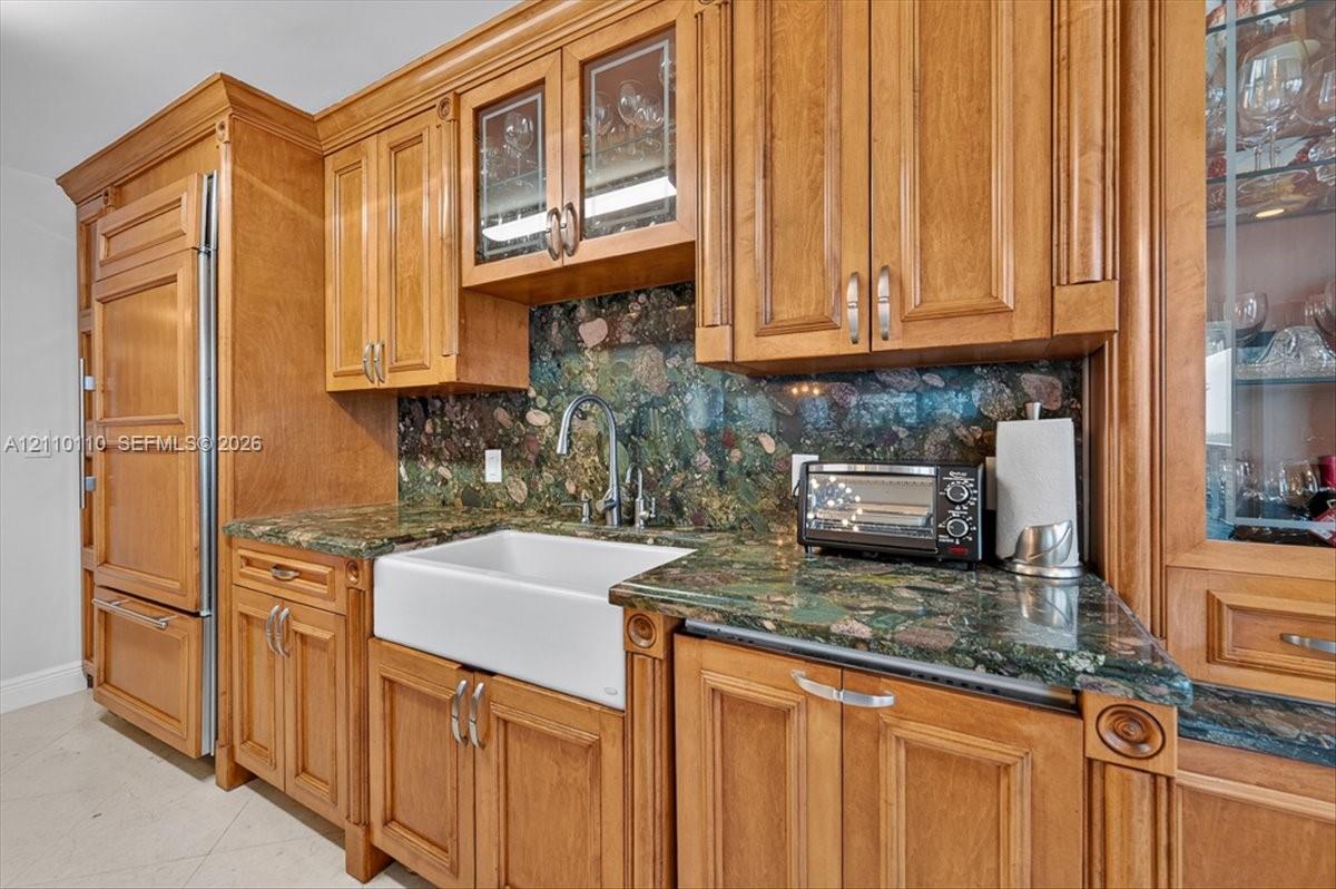 Kitchen featuring custom wood cabinetry, integrated refrigerator, stone countertops, full-height decorative stone backsplash, and a white farmhouse-style sink