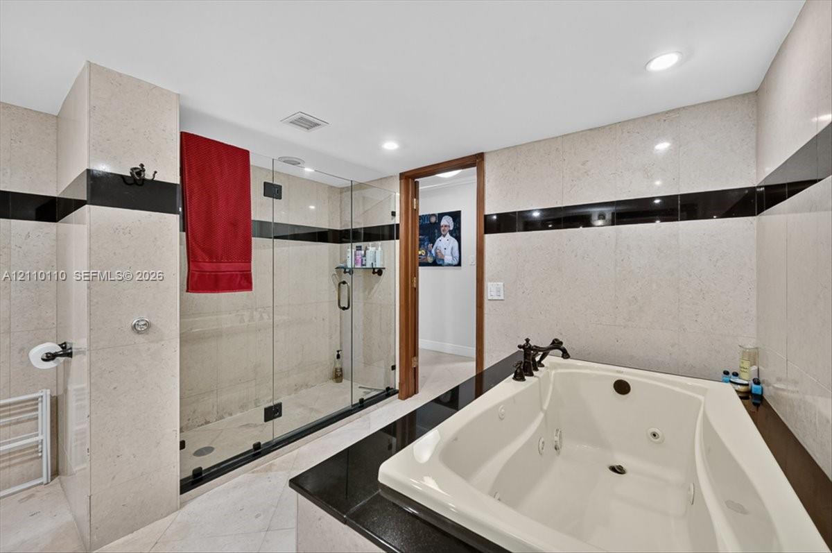 Bathroom featuring a jetted soaking tub with a black stone surround, a glass-enclosed walk-in shower, wall-to-wall light-toned tile with black inlay accents, and recessed lighting