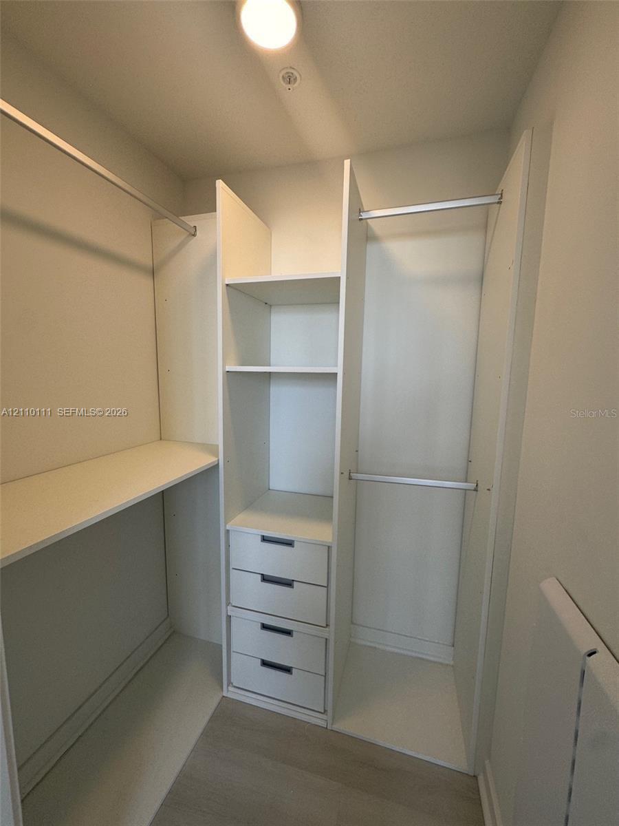 Walk-in closet featuring white built-in shelving, integrated storage drawers with matte finish hardware, dual-level hanging rods, and wood-finish flooring