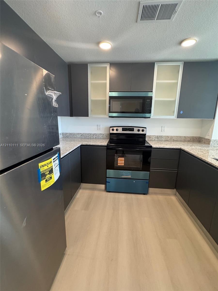 Kitchen featuring flat-panel dark cabinetry, glass-fronted upper cabinets, stone-composite countertops, stainless steel appliances, and wood-finish flooring
