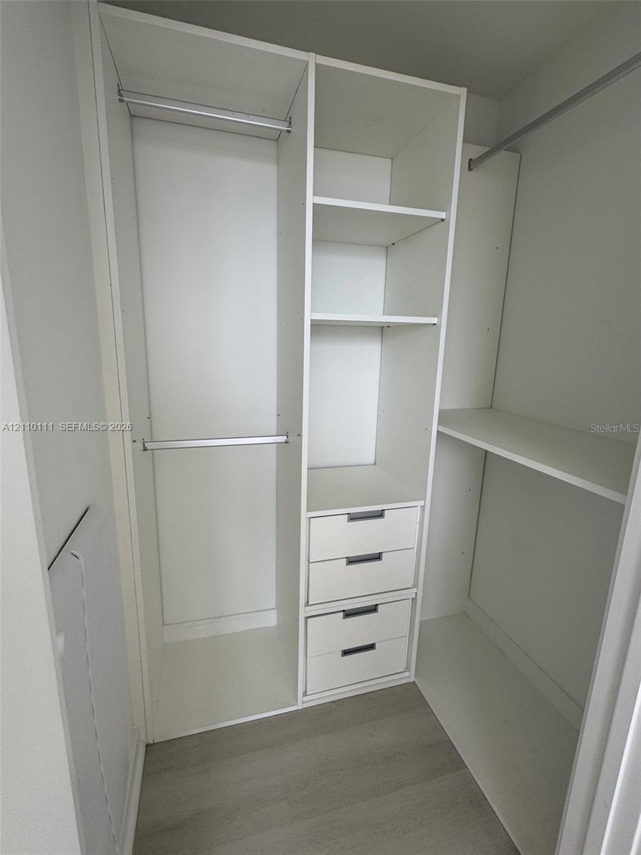 Custom walk-in closet featuring white built-in shelving, a central tower of three drawers, dual hanging rods, and wood-finish flooring