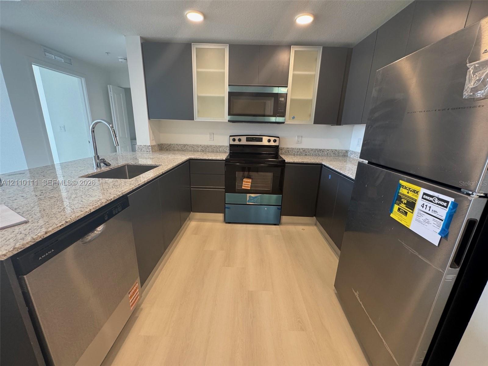 Kitchen featuring flat-panel cabinetry, light-toned stone-finish countertops, stainless steel appliances, a stainless steel undermount sink with a high-arch faucet, and light wood-finish flooring