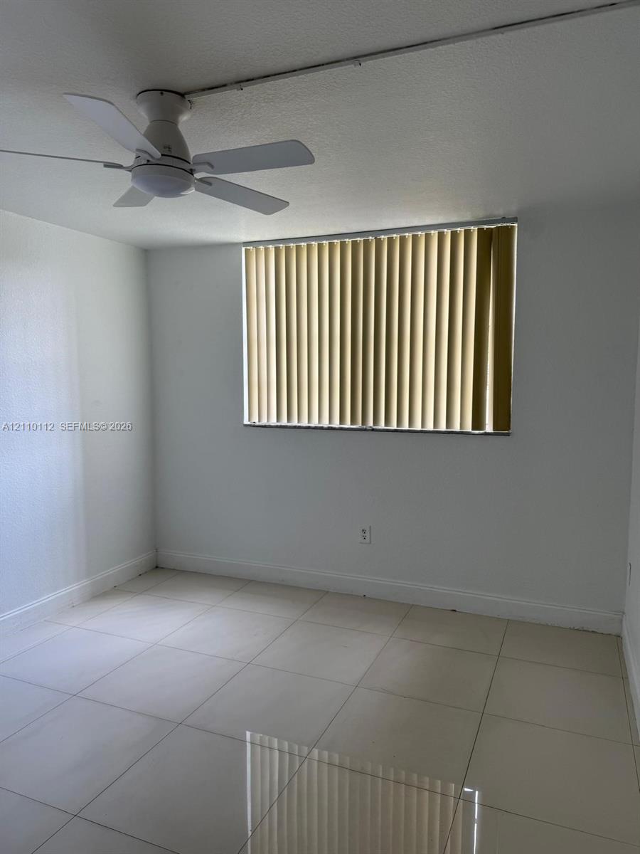 Neutral-toned tile flooring, white-painted walls, ceiling-mounted fan, and vertical window blinds