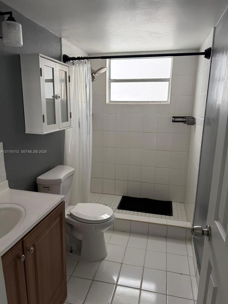 Bathroom featuring white tile flooring, a shower with white wall tile, a wood-finish vanity with a sink, and a white wall-mounted storage cabinet