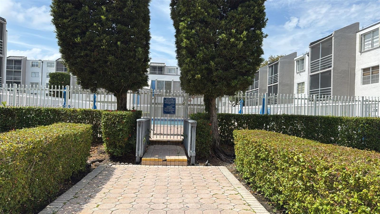 Common area swimming pool enclosed by metal fencing, featuring a paver stone walkway and mature hedgerow landscaping