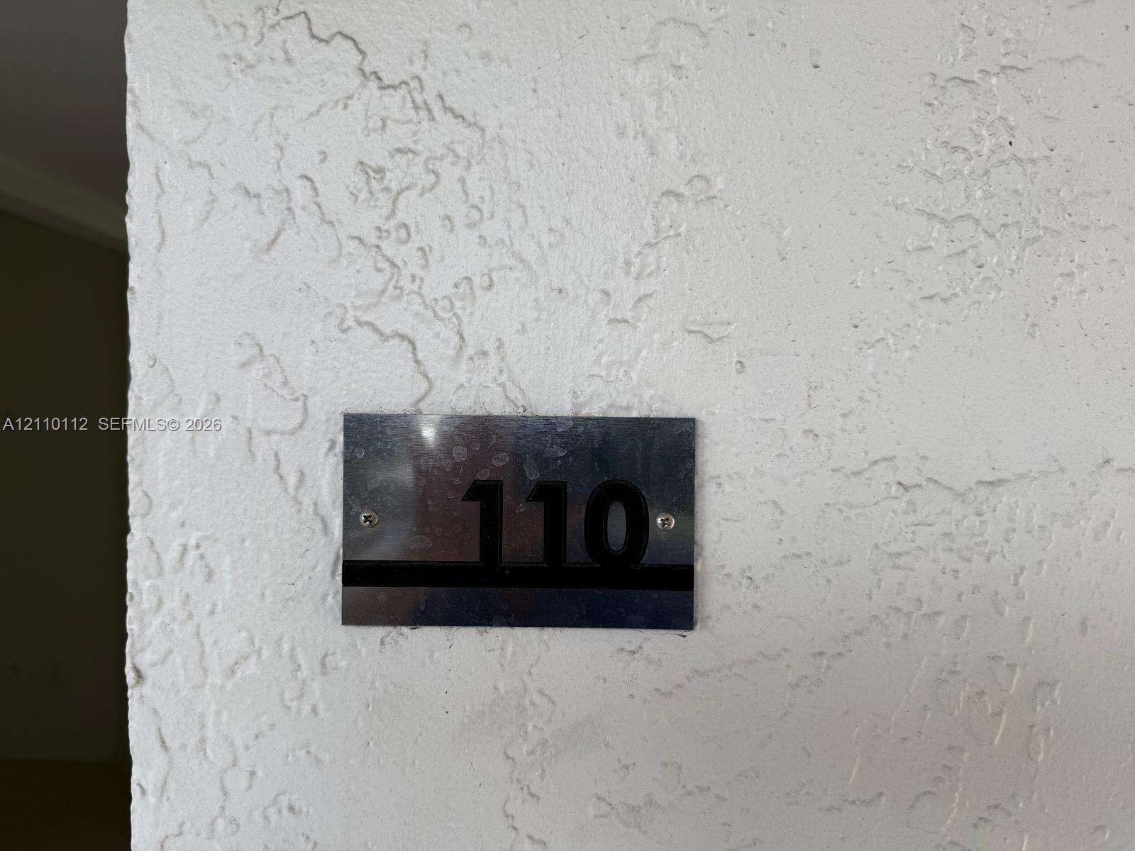 Metallic, rectangular unit identification plaque with raised black numbering mounted on a textured plaster wall surface