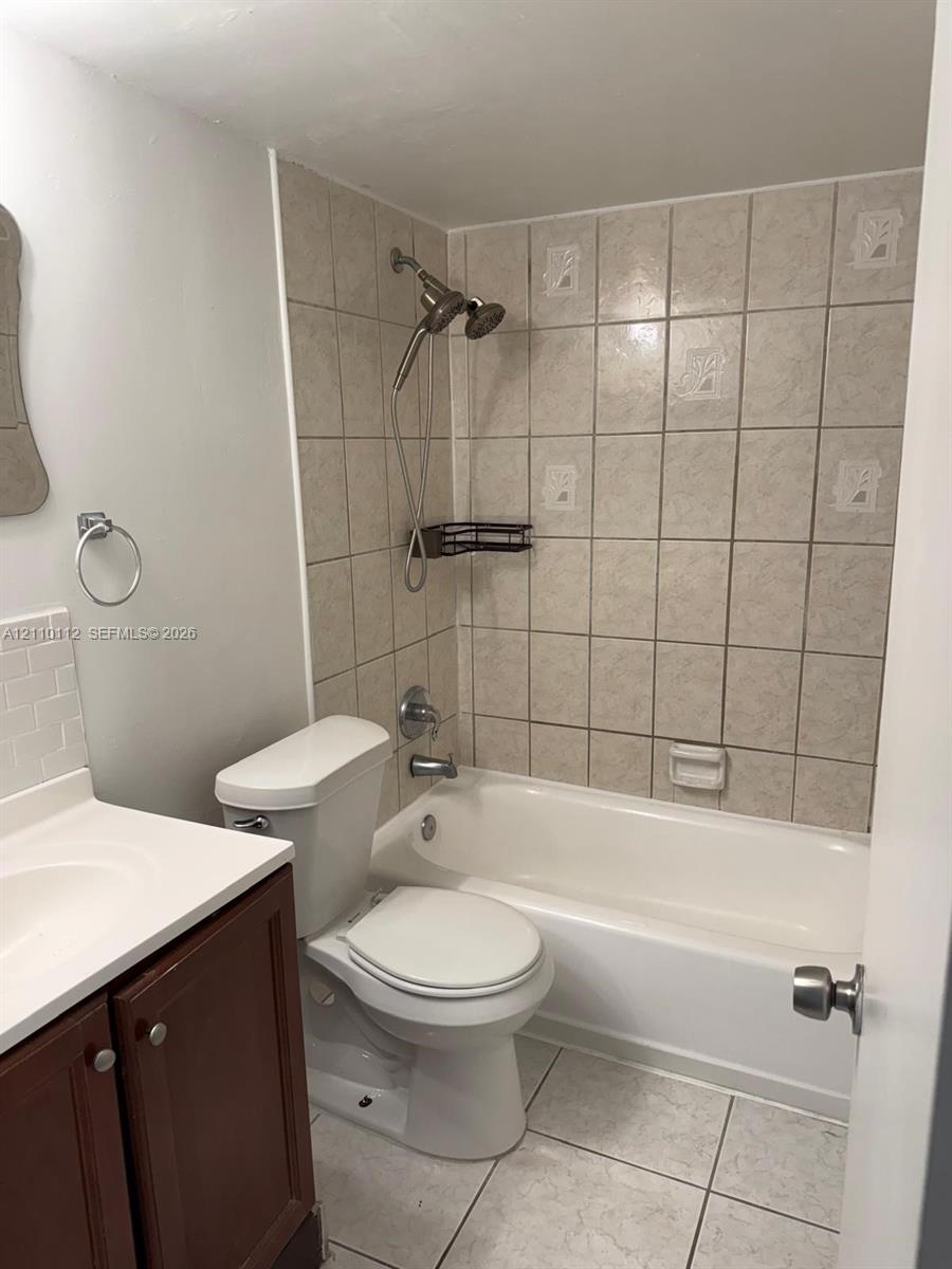 Bathroom featuring a wood-finish vanity with a white countertop, a white porcelain toilet, and a bathtub with a tiled surround and an adjustable handheld showerhead