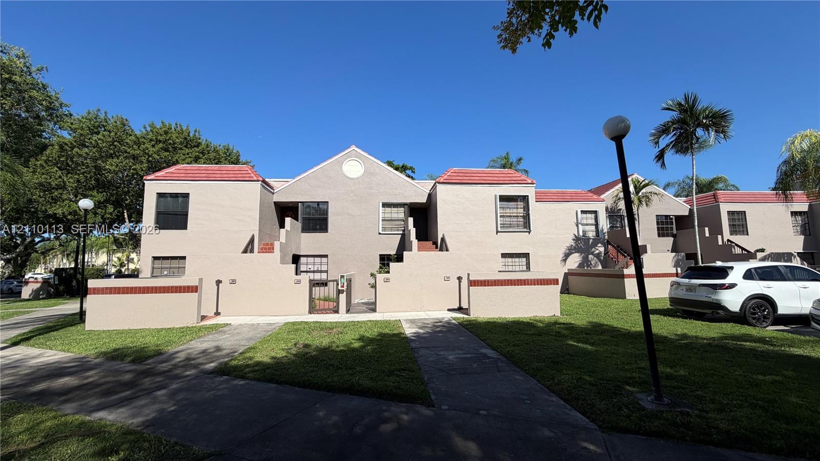 Multi-unit building exterior featuring stucco siding, red-tile roof accents, private ground-level courtyard entries, and manicured surrounding lawn space