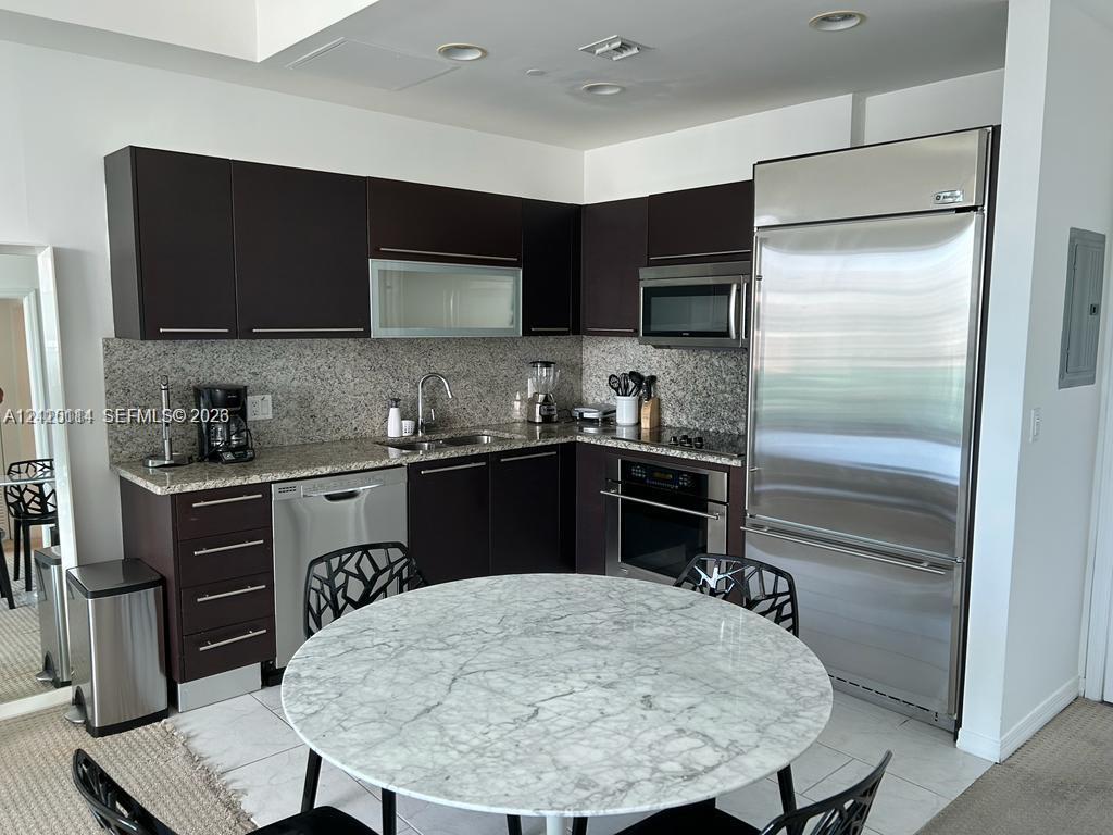 Kitchen featuring dark-toned cabinetry, light stone countertops, integrated stainless steel refrigerator, built-in oven, and recessed lighting