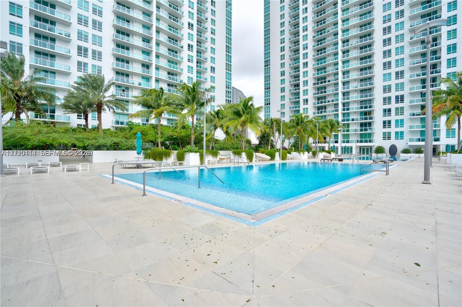 Swimming pool with metal handrails, expansive stone-tile deck, and landscaped surroundings featuring mature palm trees