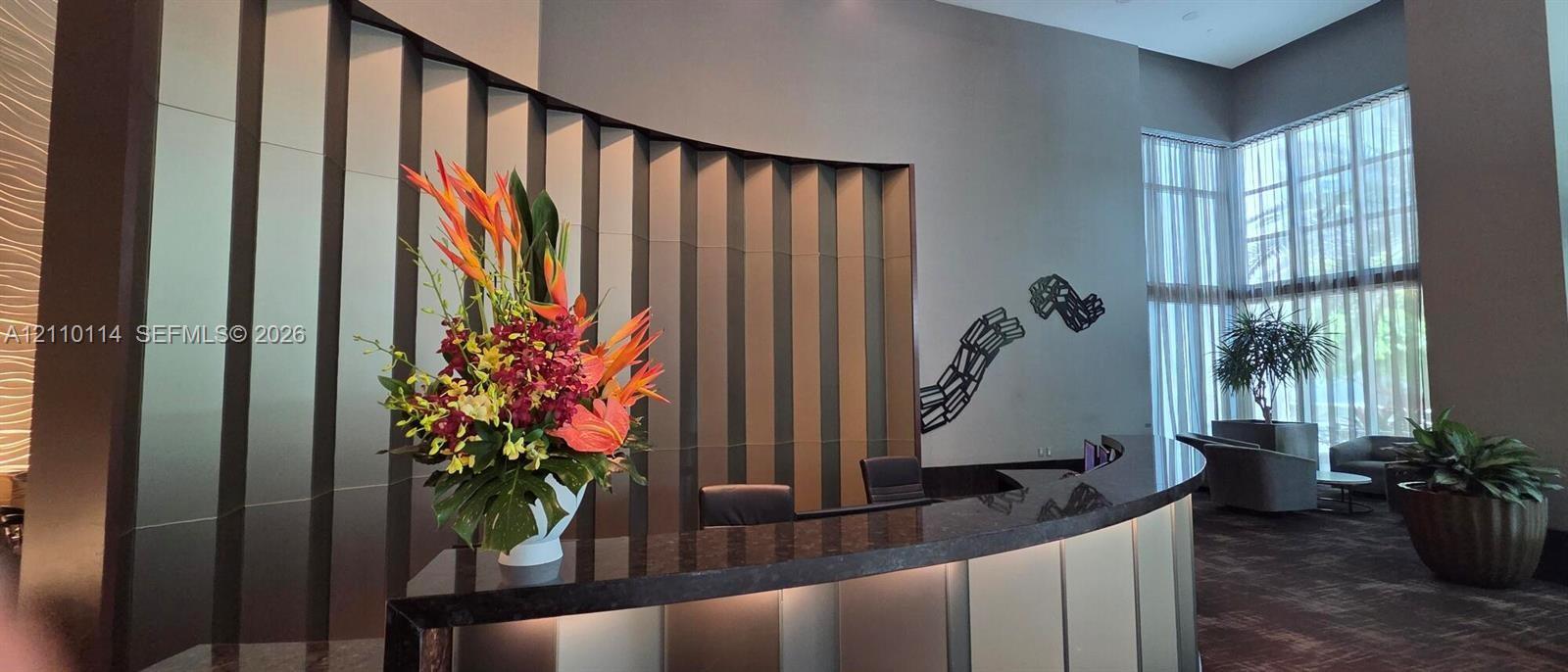 Double-height lobby featuring a curved reception desk with a stone countertop, vertical metallic wall paneling, and floor-to-ceiling windows