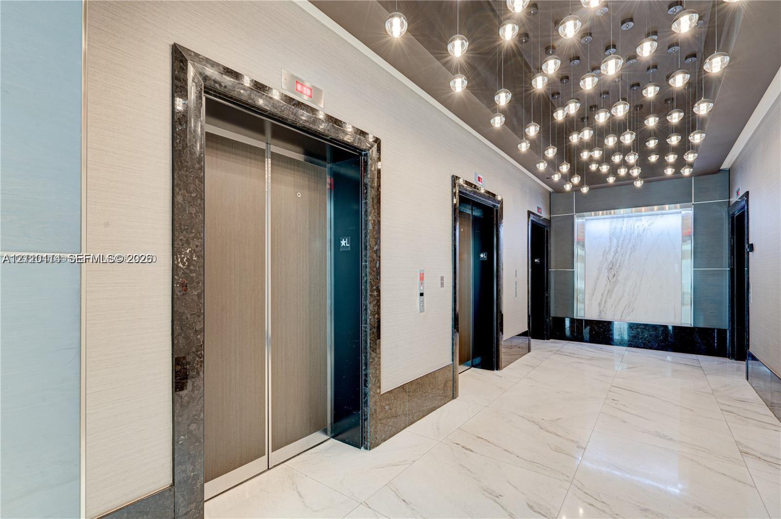 Elevator lobby featuring metallic elevator doors with stone-trim surrounds, large-format tile flooring, a decorative cluster of globe pendant lighting, and a backlit stone accent wall