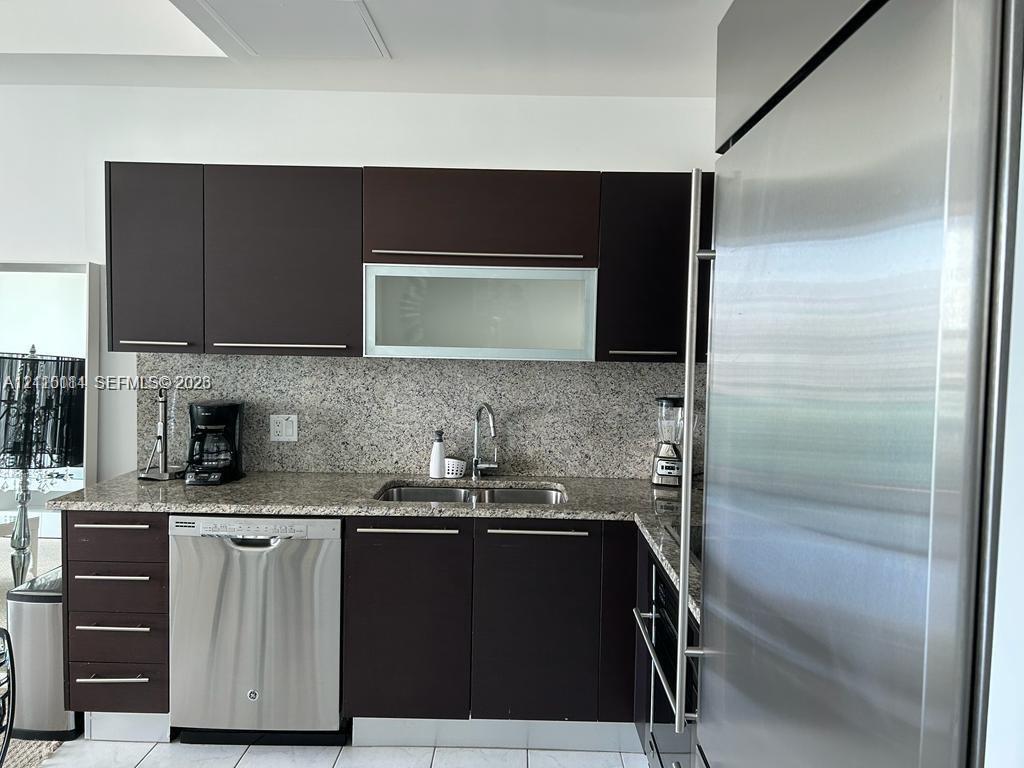 Kitchen featuring dark-finish cabinetry, stone countertop, stainless steel dishwasher, integrated stainless steel refrigerator, and undermount dual-basin sink with metallic faucet