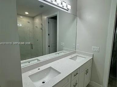 Dual-sink vanity with a solid-surface countertop, white shaker-style cabinetry, a full-width mirror, and a glass-enclosed shower with tiled surround