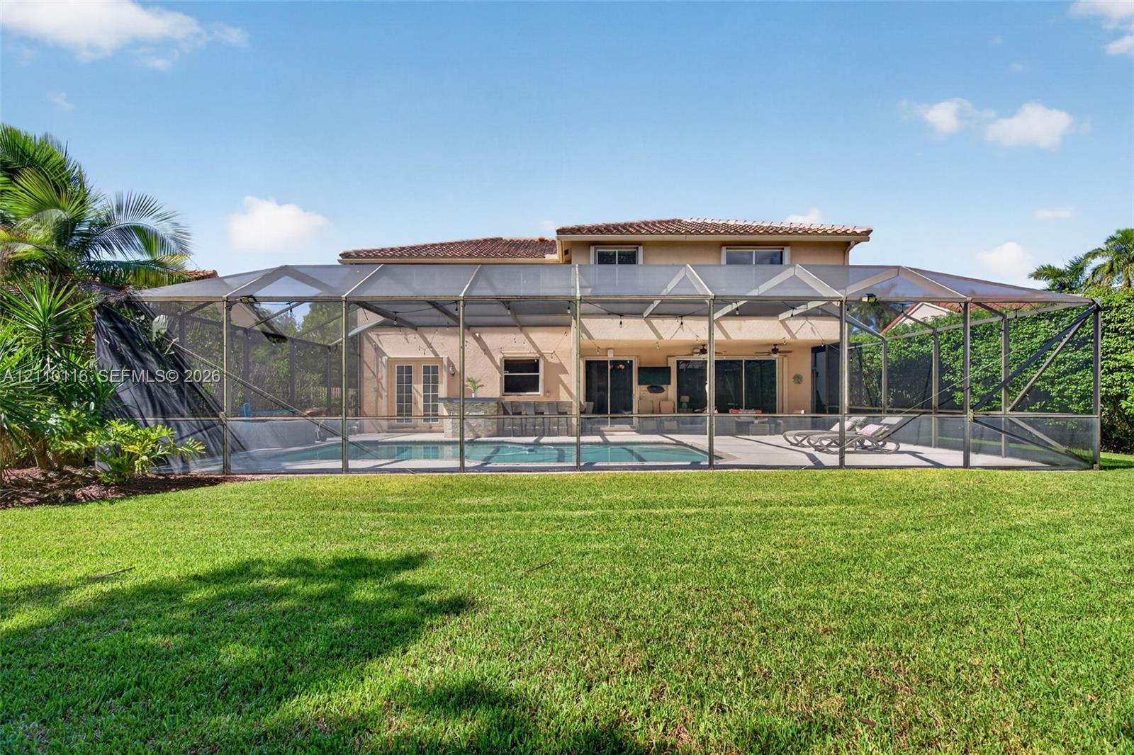Rear exterior featuring a screened-in lanai with a swimming pool, built-in outdoor kitchen, concrete patio, and a fenced grass lawn