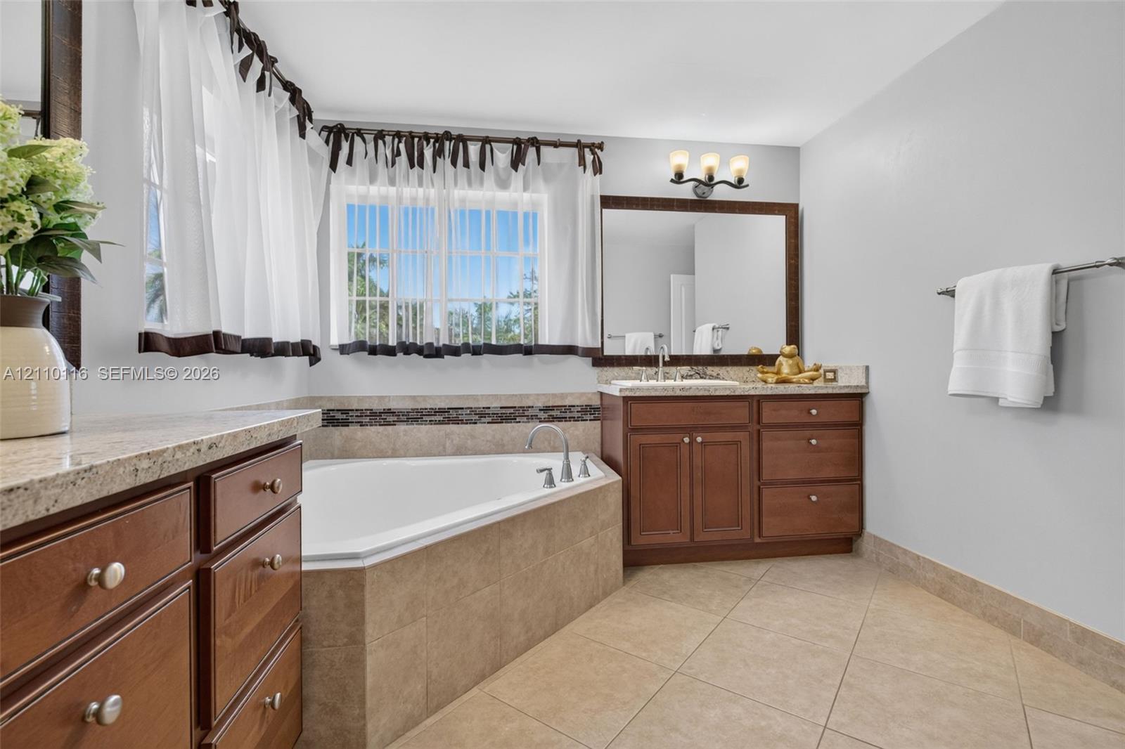Bathroom featuring a corner soaking tub with tile surround, wood cabinetry, stone-finish countertops, ceramic tile flooring, and a large framed vanity mirror