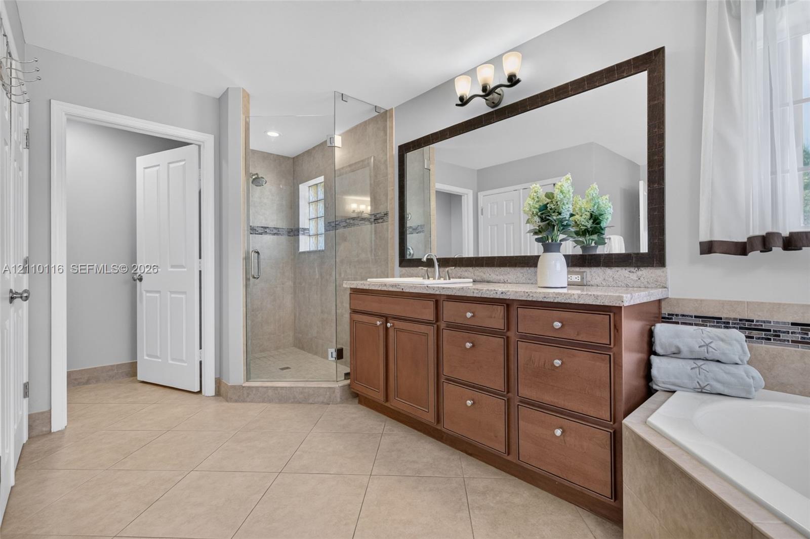 Bathroom featuring a natural wood-finish vanity with stone-surfaced countertop, glass-enclosed shower with tile surround, large framed wall mirror, and soaking tub