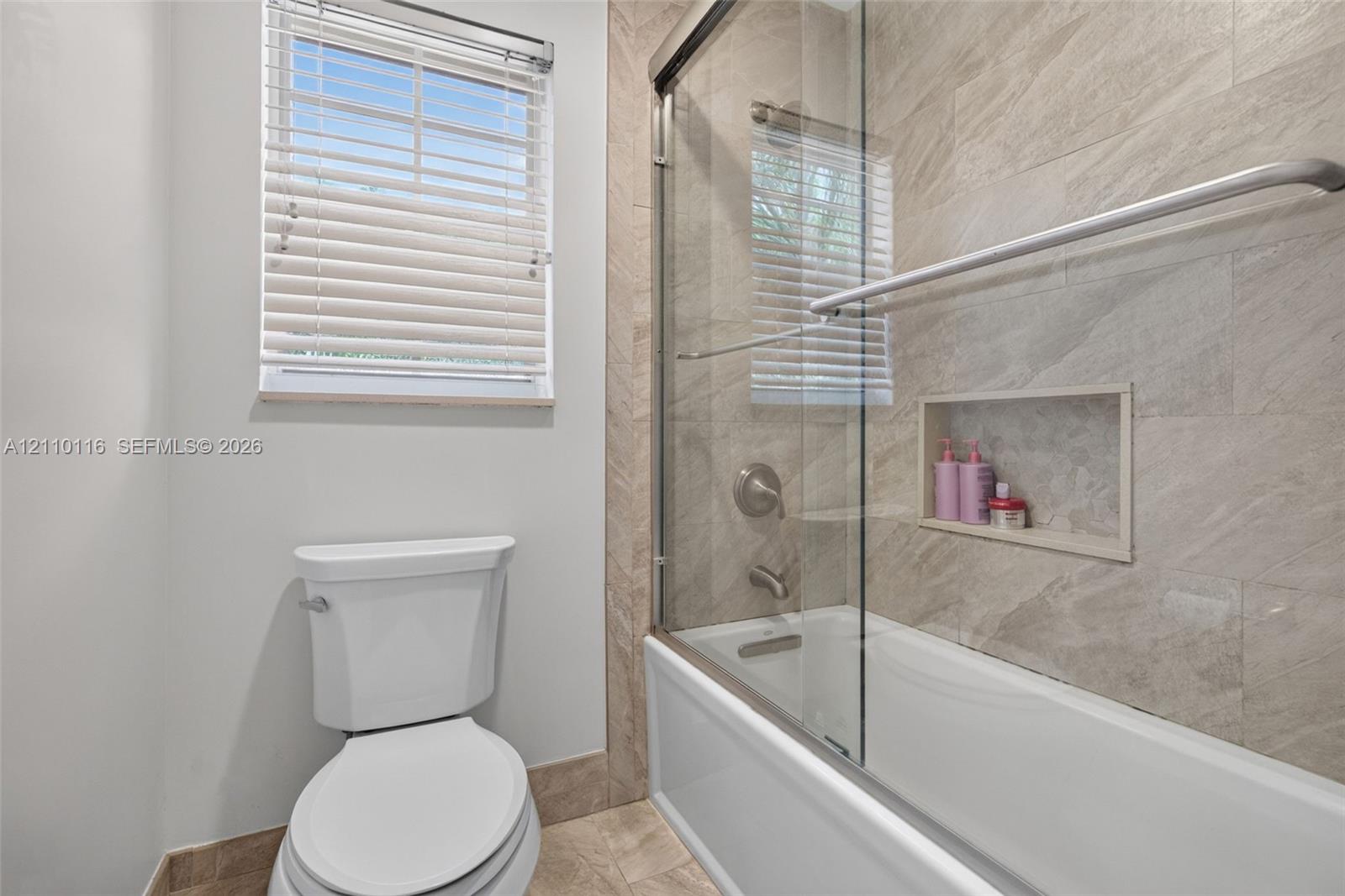 Bathroom featuring a sliding glass shower door, recessed wall niche, neutral stone-tile surround, and window with blinds
