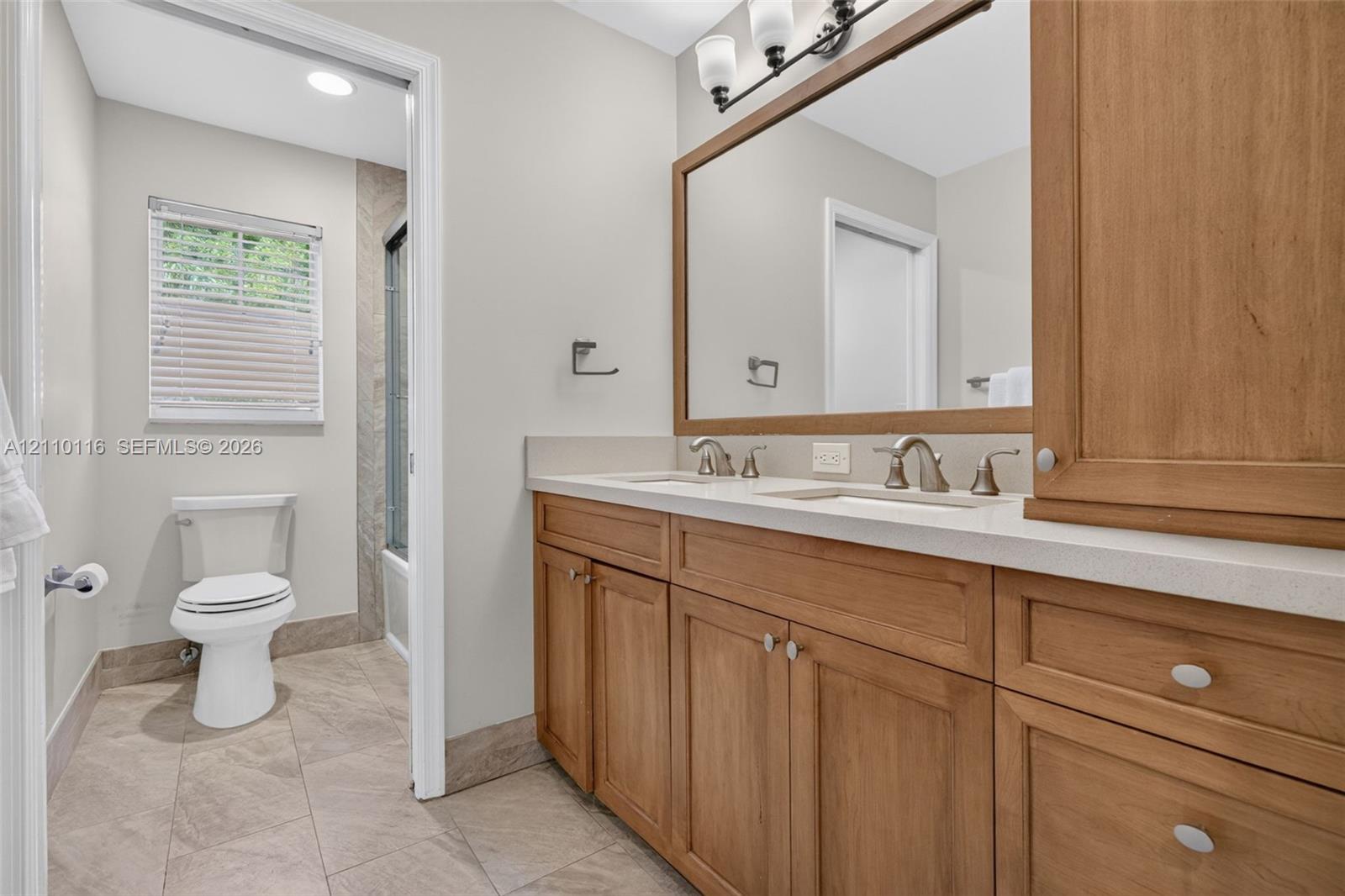Double vanity featuring wood-finish cabinetry, stone-composite countertop, dual undermount sinks, and a full-width framed mirror