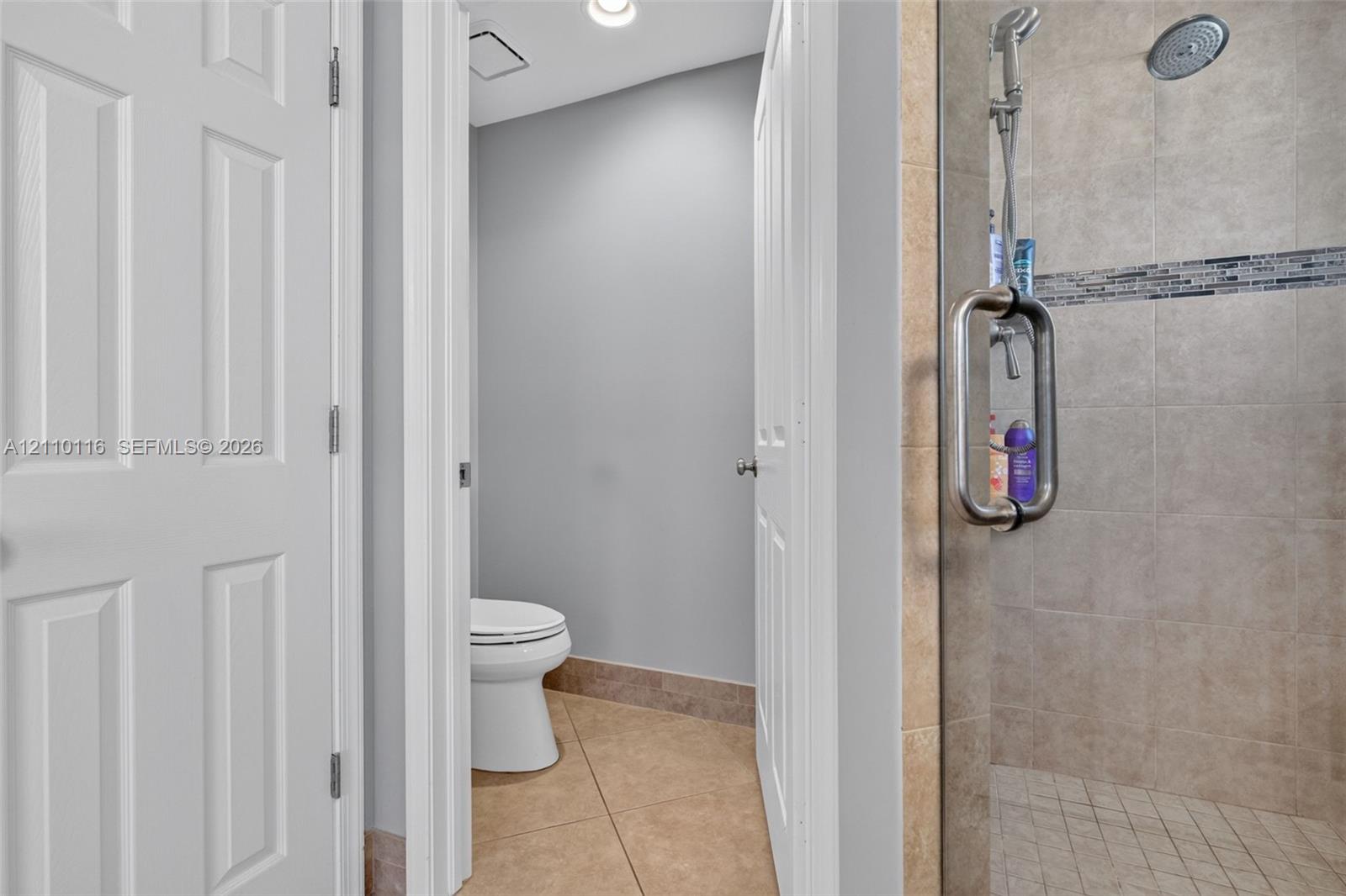 Walk-in shower with a glass door, rainfall showerhead, and tile surround featuring a decorative mosaic accent band, accompanied by tile flooring and white panel doors