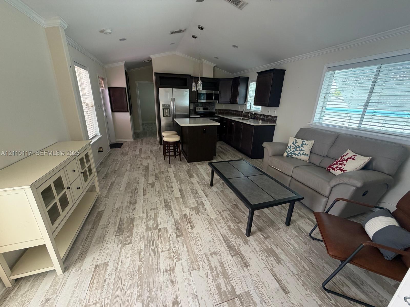 Open-concept area featuring vaulted ceilings, wood-finish flooring, dark-toned cabinetry, a central island with countertop, and stainless steel kitchen appliances