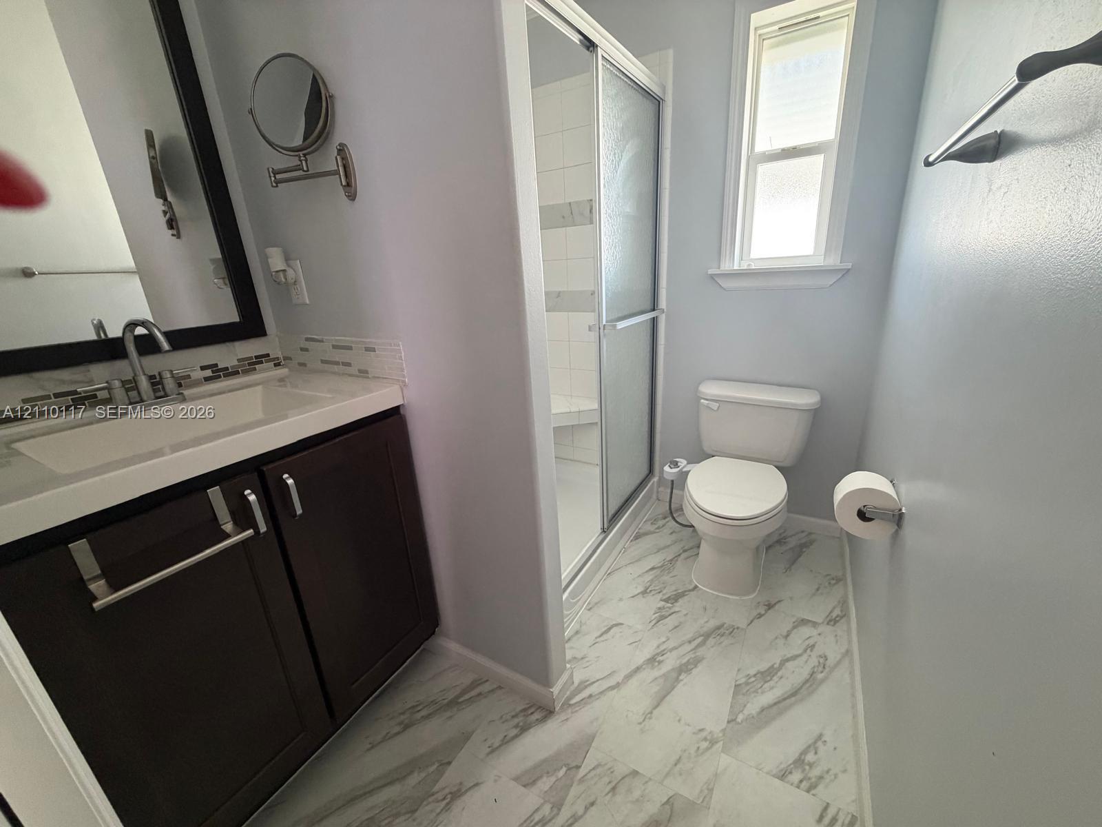 Bathroom featuring a dark-finish vanity with a rectangular sink, stone-pattern flooring, a walk-in shower with a glass enclosure, and a window