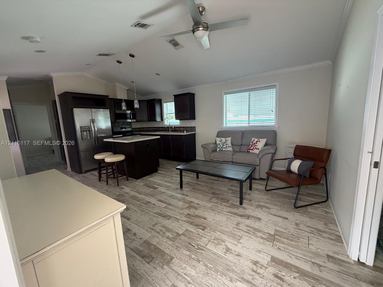 Open-concept interior featuring wood-finish flooring, dark-toned kitchen cabinetry, stainless steel refrigerator, kitchen island, and recessed ceiling lighting