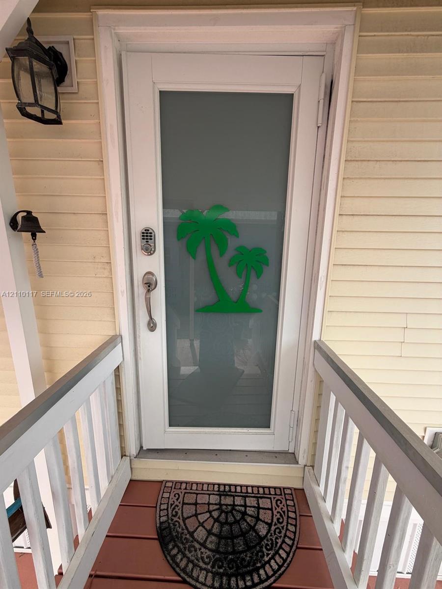 Entry door featuring an integrated glass panel with decorative palm tree motif, electronic keypad lock, brushed metal hardware, and exterior wall-mounted carriage-style lighting Entry door featuring an integrated glass panel with decorative palm tree motif, electronic keypad lock, brushed metal hardware, and exterior wall-mounted carriage-style lighting