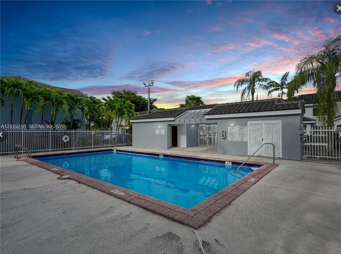 Rectangular swimming pool featuring a brick paver border, concrete decking, metal perimeter fencing, and an attached auxiliary building with a tiled roof