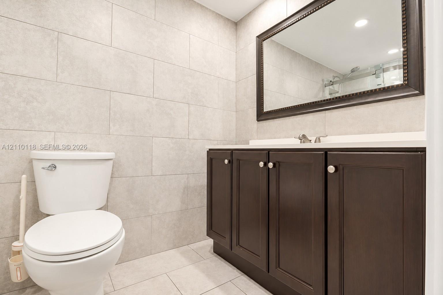 Bathroom featuring large-format wall and floor tiling, dark wood-finish cabinetry, a solid surface countertop, and a framed mirror