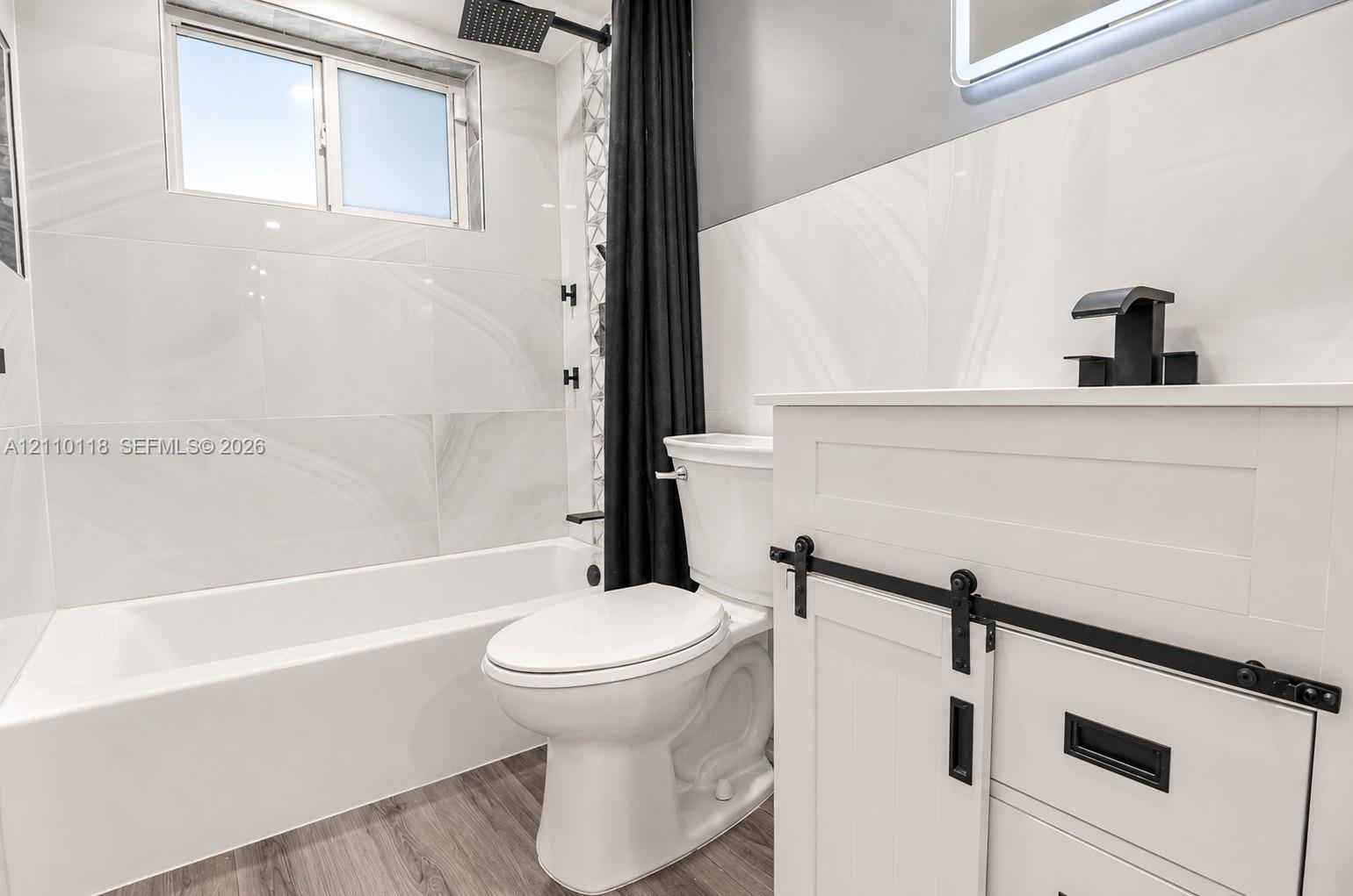 Bathroom featuring large-format wall tile, wood-finish flooring, a white vanity with matte black hardware, and a rainfall showerhead