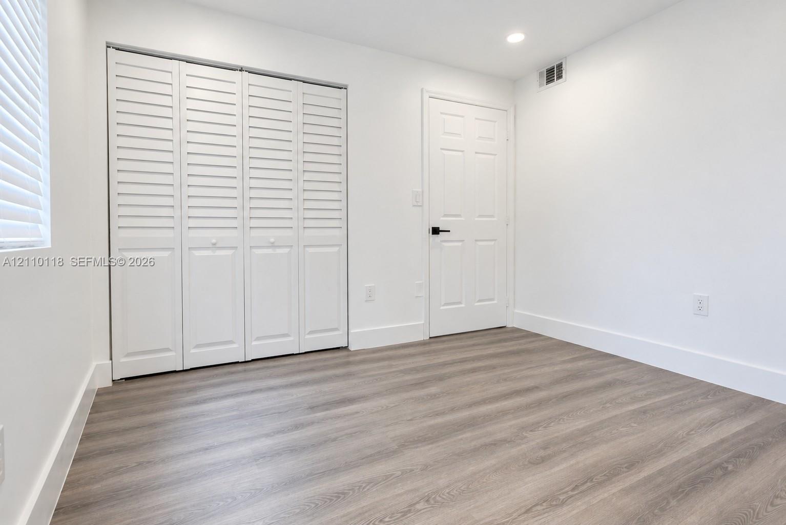 Neutral interior featuring wood-finish flooring, white louvered closet doors, a six-panel door with matte black hardware, and recessed lighting