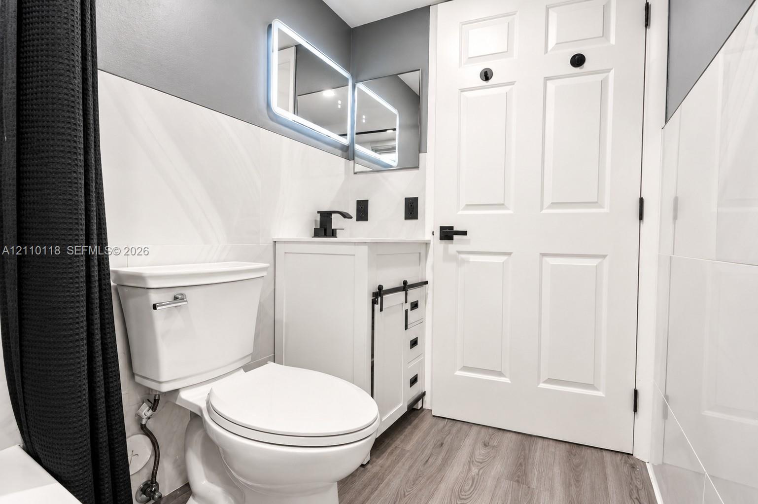 Bathroom featuring a white vanity cabinet with black sliding barn-door hardware, an integrated sink with a matte black faucet, a rectangular LED-backlit mirror, and wood-finish flooring