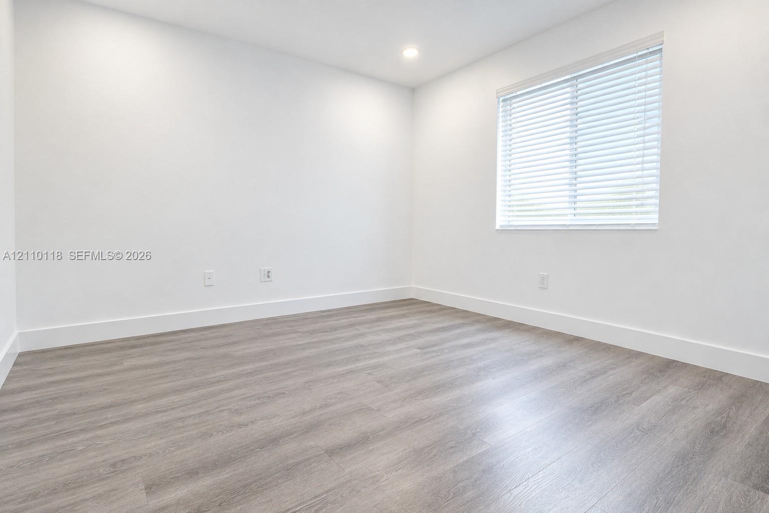 Neutral interior space featuring wood-finish flooring, crisp white walls, recessed ceiling lighting, and a window with horizontal blinds