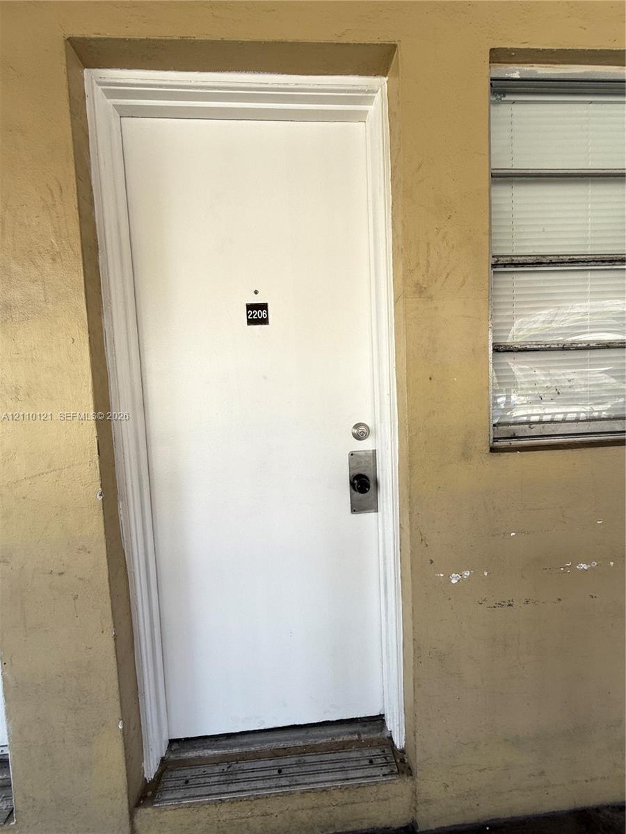 Exterior solid-core door featuring a brushed metal deadbolt assembly, integrated door viewer, and white finish, complemented by a horizontal jalousie window with metal frames