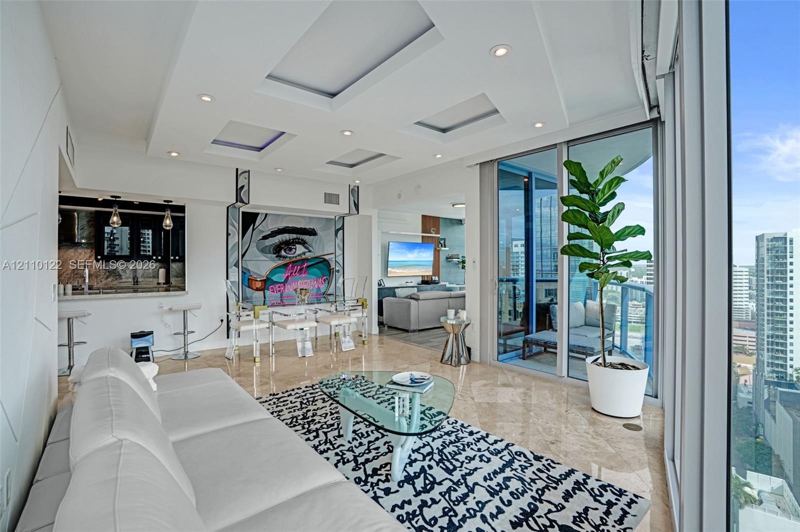 Living area featuring floor-to-ceiling glass sliding doors, tray ceiling with recessed lighting, polished stone flooring, and a kitchen pass-through window with an integrated breakfast bar