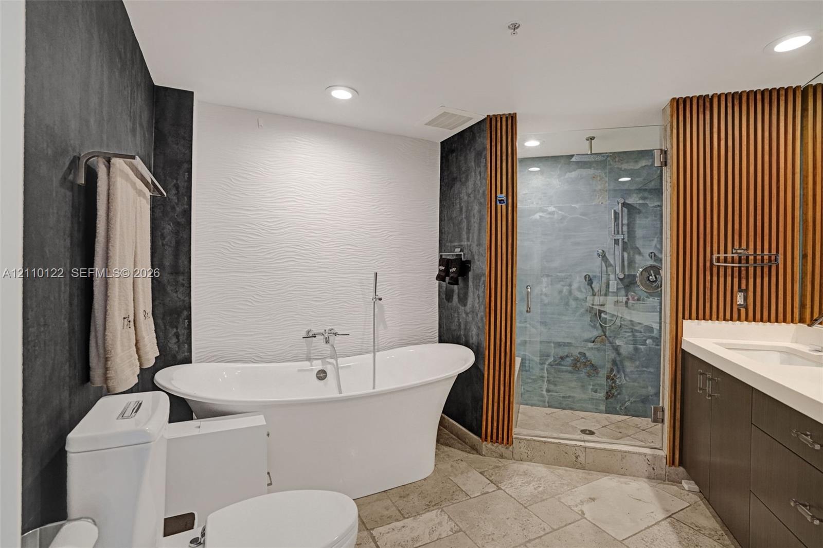 Freestanding soaking tub with a floor-mounted faucet, walk-in glass shower with stone tile interior, vertical wood-slat accent wall, stone-finish floor tiling, and dark-toned cabinetry with a solid-surface vanity countertop
