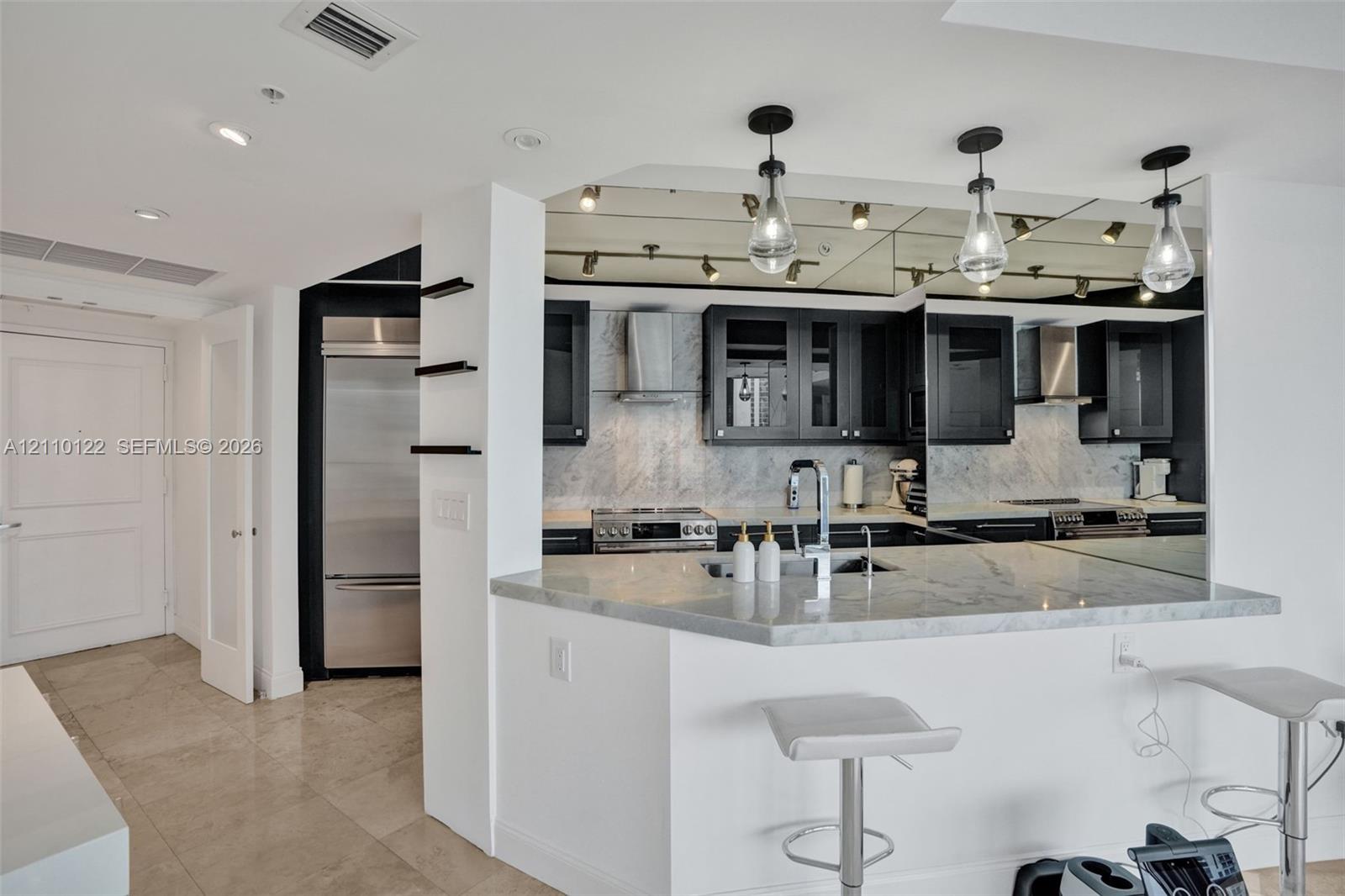 Kitchen featuring stone-surfaced countertops, custom cabinetry with glass-front accents, stainless steel appliances, mirrored backsplash, and pendant lighting