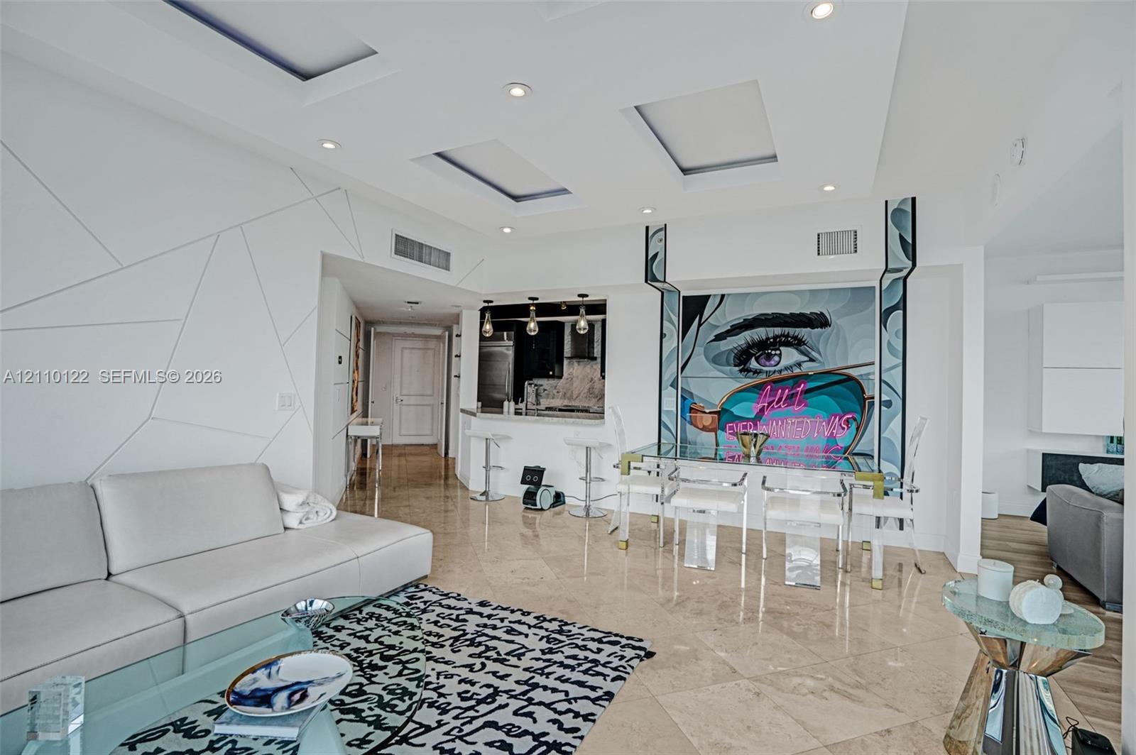 Open-concept living space featuring stone-finish flooring, recessed ceiling lighting with geometric coffers, a pass-through kitchen bar, and a wall accent with integrated lighting